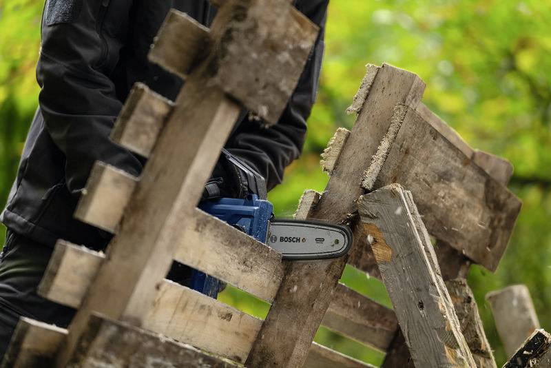Eine Person zersägt mit einer Motorsäge aus Holzpaletten im Freien. Hintergrund zeigt grüne Vegetation.