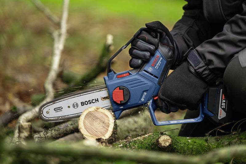 Eine Person sägt mit einer blauen Bosch-Akkukettensäge einen Holzstamm auf einer grünen Wiese.