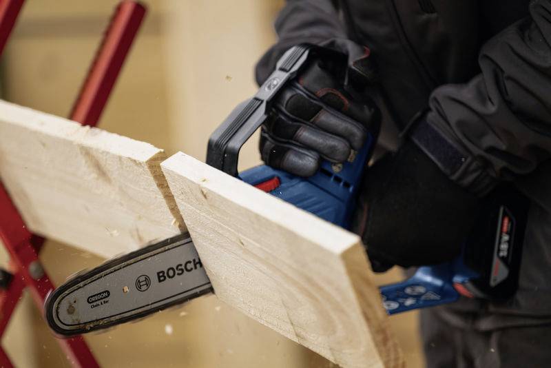 Person arbeitet mit einer batteriebetriebenen Kettensäge, um ein Holzbrett zu zersägen. Fokus auf Sicherheitshandschuhe und Werkzeug.