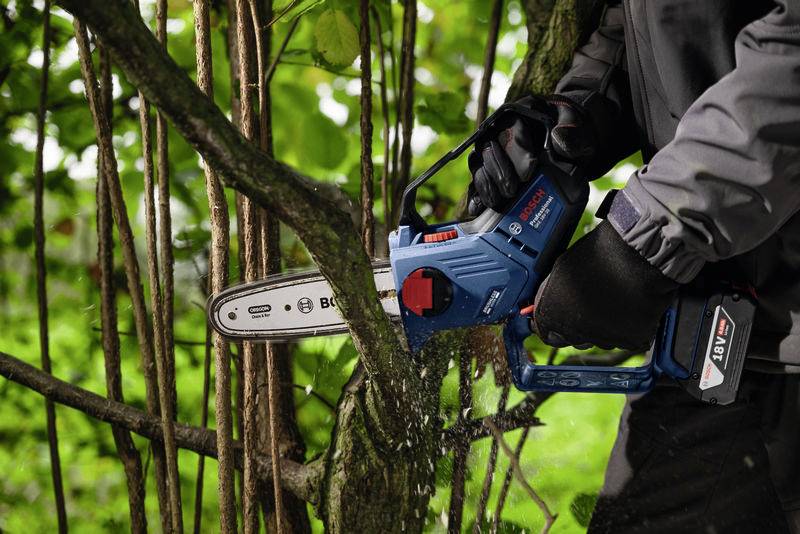 Ein Mensch sägt mit einer blauen Motorsäge Äste im Wald. Es fliegen Sägespäne durch die Luft. Die Person trägt eine dunkle Jacke und Handschuhe.