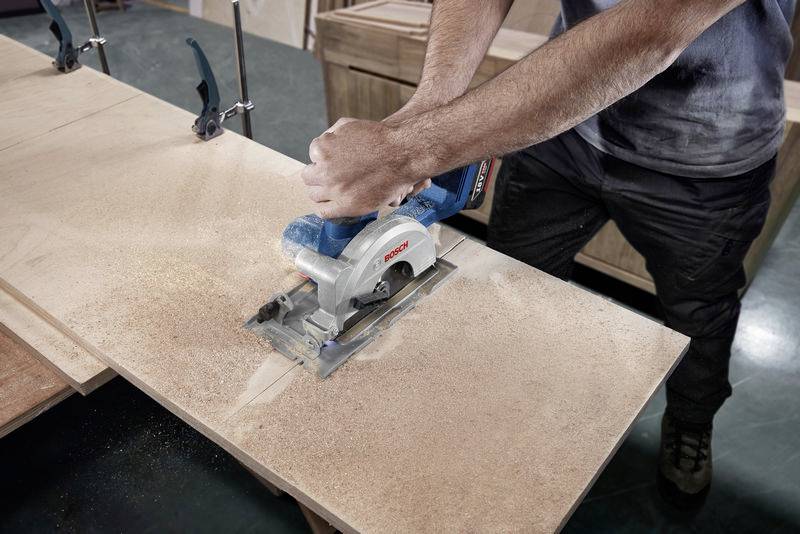 Eine Person sägt mit einer elektrischen Handkreissäge ein großes Holzbrett. Späne fliegen zur Seite. Holzwerkstatt im Hintergrund.