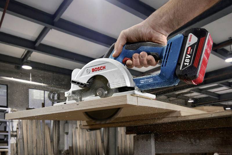 Eine Hand führt eine Bosch-Akkukreissäge über ein Holzbrett in einer Werkstatt. Staubpartikel wirbeln während des Schneidens auf.