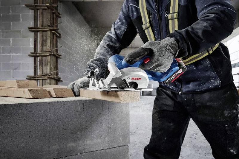Eine Person sägt mit einer elektrischen Säge ein Holzbrett auf einer Baustelle. Im Hintergrund sind Wände aus Betonblöcken erkennbar.