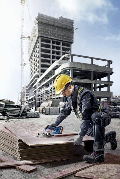 Ein Bauarbeiter mit Helm schneidet Holzplatten auf einer Baustelle. Im Hintergrund ist ein im Bau befindliches Gebäude zu sehen.