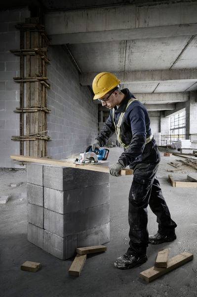 Ein Bauarbeiter mit Helm und Schutzkleidung nutzt eine Handkreissäge, um ein Holzbrett in einem unfertigen Gebäude auf Betonblöcken zu schneiden.