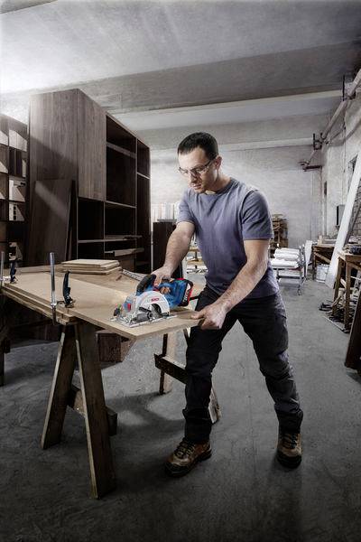 Ein Mann sägt eine Holzplatte mit einer elektrischen Handkreissäge in einer Werkstatt, umgeben von Regalen und Holzmaterialien.