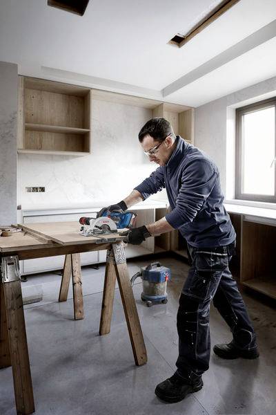 Eine Person arbeitet in einer Werkstatt an einem Holztisch mit einer elektrischen Säge. Im Hintergrund sind Regale und ein Fenster zu sehen.
