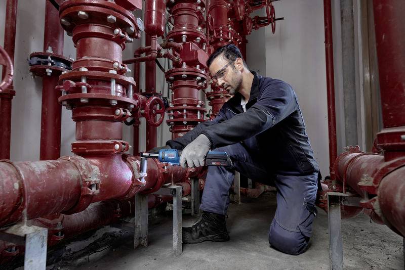 Ein Handwerker in Arbeitskleidung kniet und arbeitet mit einem Elektrowerkzeug an einem roten Rohrsystem in einem technischen Raum.