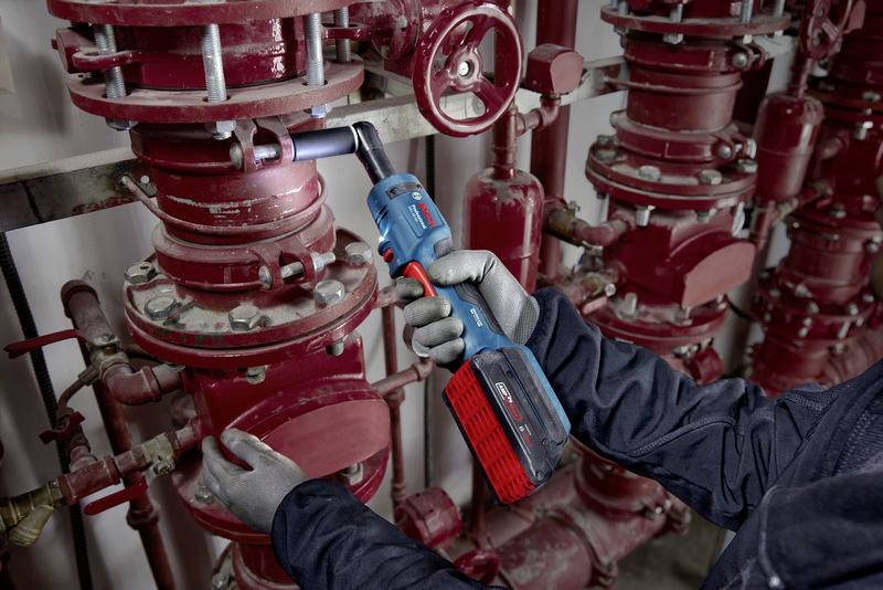 Eine Person fixiert mit einem Akkuschrauber ein rotes Ventil in einer industriellen Rohrleitungsanlage, umgeben von mehreren Rohren.