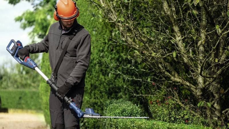 Ein Gärtner mit Schutzkleidung schneidet eine Hecke mit einer elektrischen Heckenschere in einem grünen Garten.