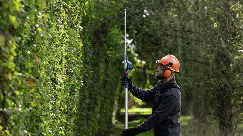Ein Gärtner mit Schutzhelm und Gehörschutz schneidet eine hohe Hecke mit einer langen Heckenschere in einem Garten.