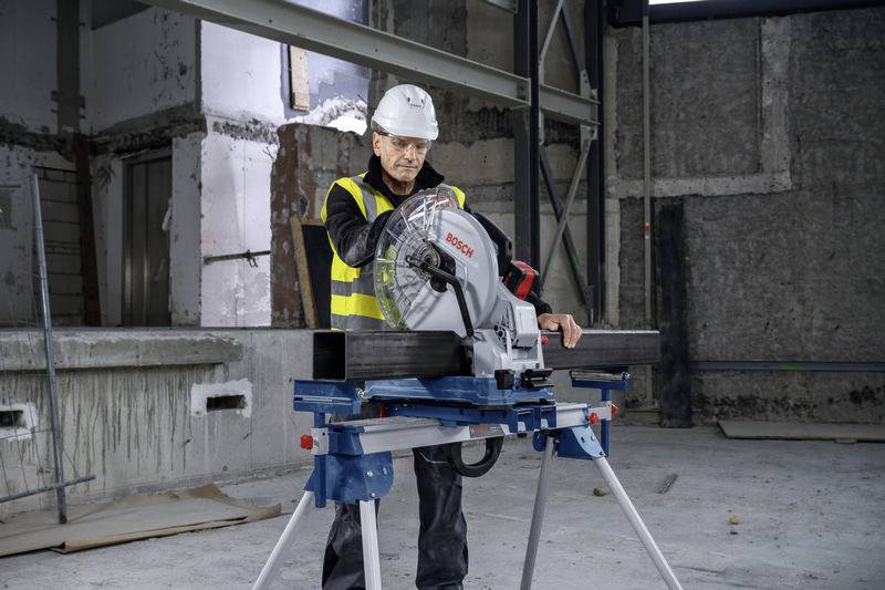 Ein Bauarbeiter mit Schutzhelm und Warnweste schneidet mit einer Kappsäge ein Metallrohr in einer unfertigen Betonumgebung.