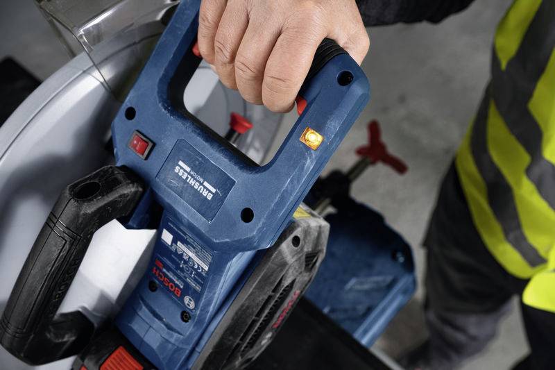 Eine Person hält ein elektrisches Werkzeug mit einem blauen Gehäuse, vermutlich eine Säge, in der rechten Hand. Sie trägt Schutzkleidung.