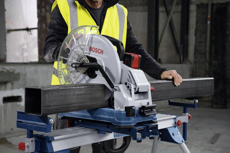 Eine Person schneidet ein Metallrohr mit einer Bosch-Kappsäge. Sie trägt eine gelbe Sicherheitsweste in einer Werkstattumgebung.