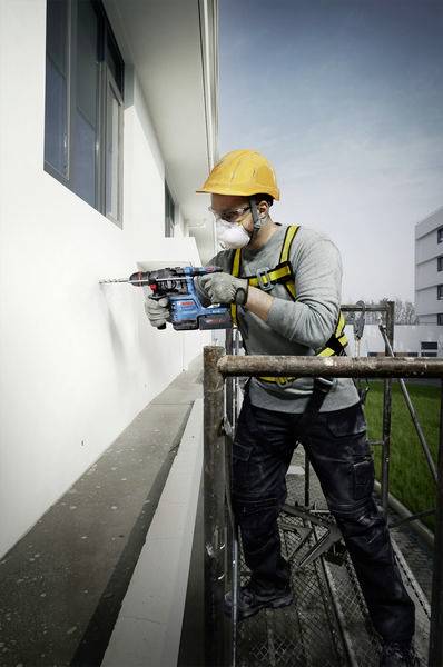 Ein Bauarbeiter mit gelbem Helm und Maske benutzt eine Bohrmaschine an einer weißen Hauswand, stehend auf einem Gerüst.