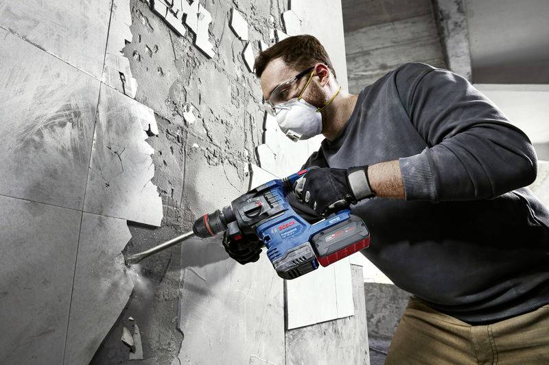 Ein Mann bohrt mit einer elektrischen Bohrmaschine ein Loch in eine Wand. Er trägt Schutzbrille und Atemmaske in industrieller Umgebung.