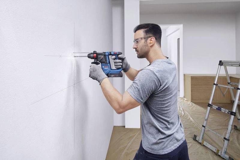 Ein Mann mit Brille bohrt mit einer Handbohrmaschine in eine weiße Wand. Er trägt graue T-Shirts und Handschuhe.