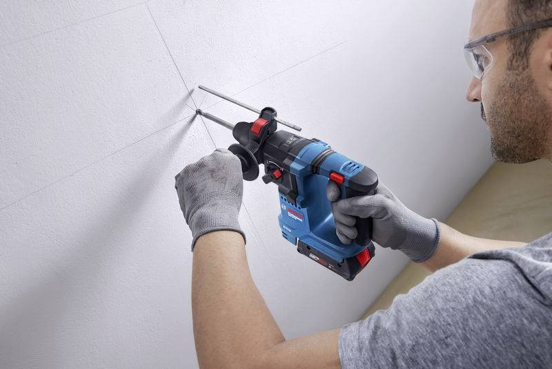 Ein Mann bohrt mit einem blauen, roten und schwarzen Bohrhammer in eine weiße Wand, trägt Schutzbrille und Handschuhe.