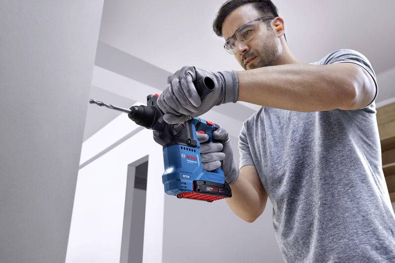 Ein Mann benutzt eine blaue elektrische Bohrmaschine, um in eine Wand zu bohren. Er trägt Schutzbrille und Arbeitshandschuhe.
