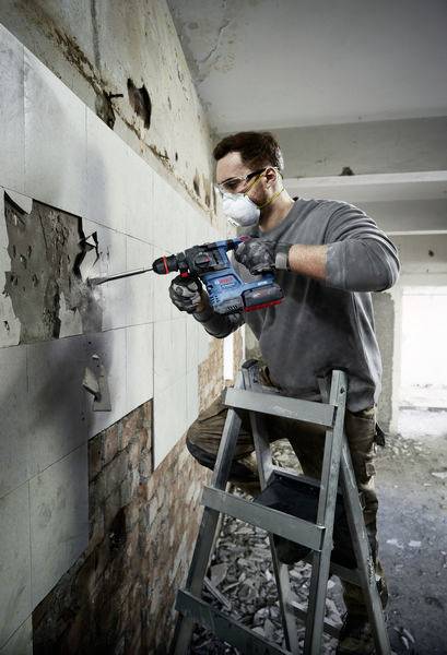 Ein Mann auf einer Leiter entfernt Fliesen von einer Wand mit einem Bohrhammer in einem renovierungsbedürftigen Raum.