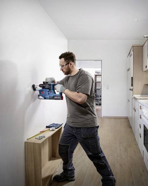 Ein Mann bohrt ein Loch in die Wand einer modernen Küche. Er trägt Schutzbrille und Handschuhe. Werkzeug liegt auf dem Regal neben ihm.