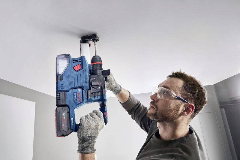 Eine Person bohrt mit einem blauen Bohrhammer in eine Decke. Sie trägt Schutzbrille und Handschuhe.