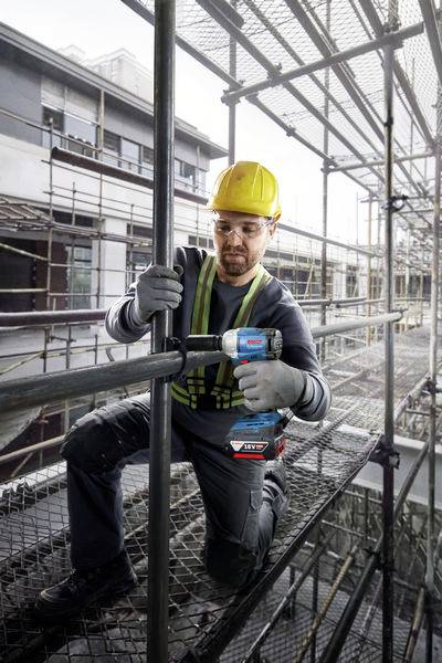 Ein Arbeiter in Sicherheitskleidung und Helm befestigt ein Gerüst mit einem Akkuschrauber. Hintergrund zeigt Baustelle und Gebäude.