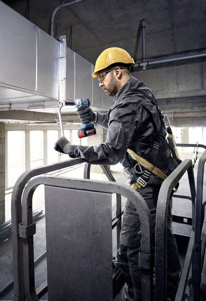 Ein Bauarbeiter mit Schutzhelm und Sicherheitsgurt steht auf einer Hebebühne und verwendet ein Elektrowerkzeug, um Leitungen zu installieren.