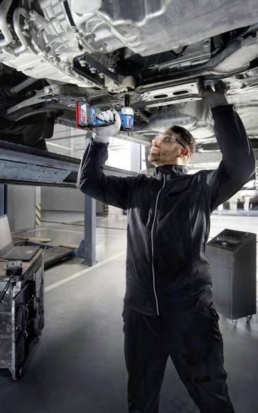 Mechaniker in Werkstatt arbeitet mit Elektrowerkzeug an der Unterseite eines angehobenen Fahrzeugs.