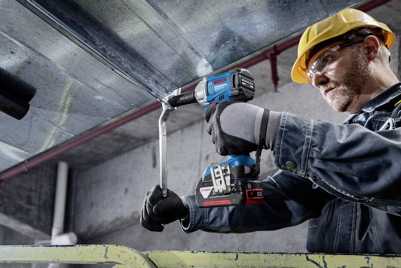 Ein Bauarbeiter mit Schutzhelm schraubt mit einem Akkuschrauber an einem Metallträger in einem industriellen Umfeld.