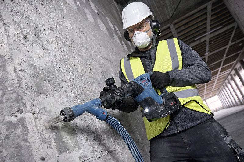 Ein Bauarbeiter trägt Schutzkleidung, benutzt einen Bohrhammer, um in eine Betonwand zu bohren. Sicherheit und Präzision am Arbeitsplatz.