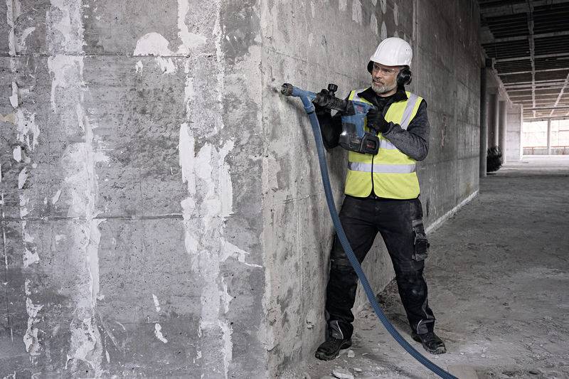 Ein Bauarbeiter in Sicherheitskleidung und Helm benutzt eine Bohrmaschine, um Löcher in eine Betonwand zu bohren.