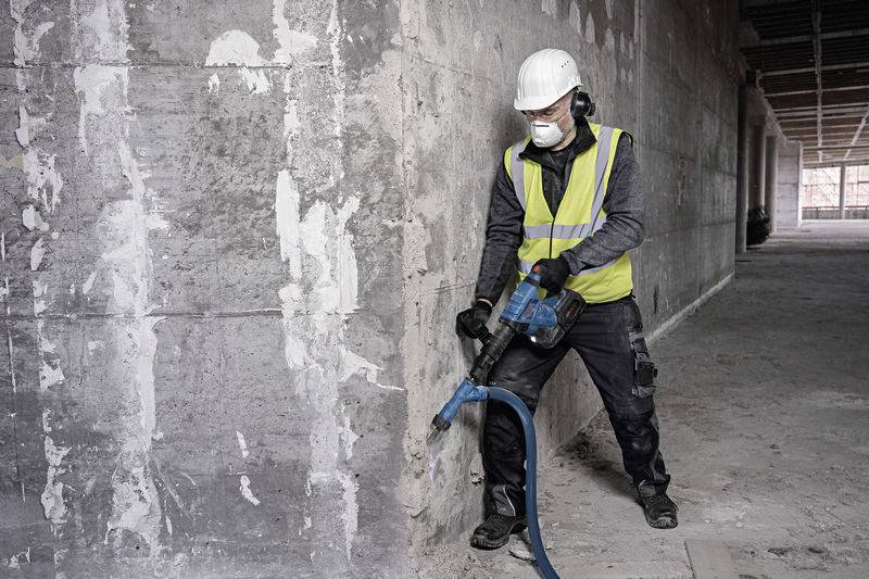 Ein Arbeiter in Sicherheitskleidung verwendet einen Bohrhammer, um eine Betonwand in einem unfertigen Gebäude abzutragen.