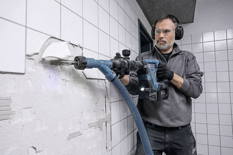 Ein Mann trägt Schutzbrille und Gehörschutz, während er mit einer Bohrmaschine Fliesen von einer Wand entfernt.
