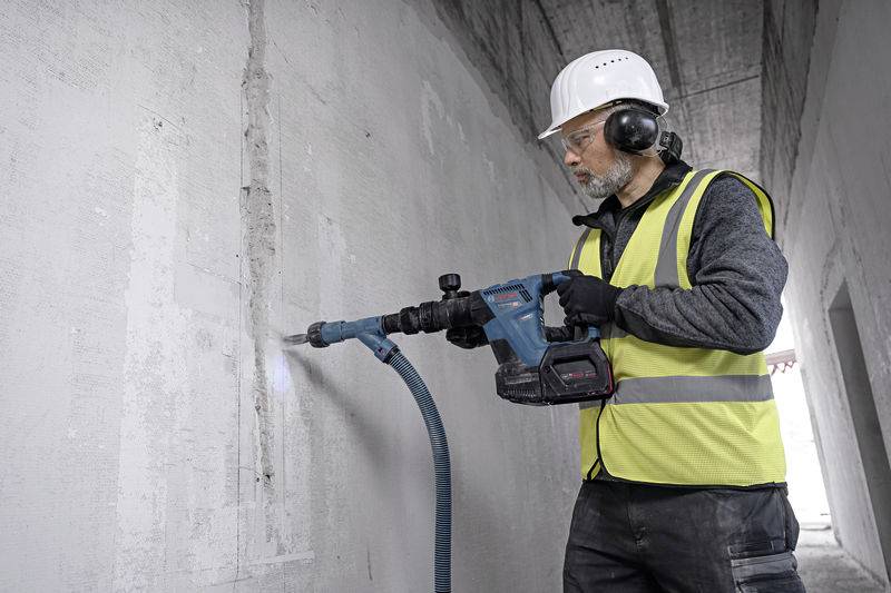 Ein Bauarbeiter trägt Helm und Schutzkleidung und benutzt einen Elektromeißel, um in einer Betonwand zu bohren.