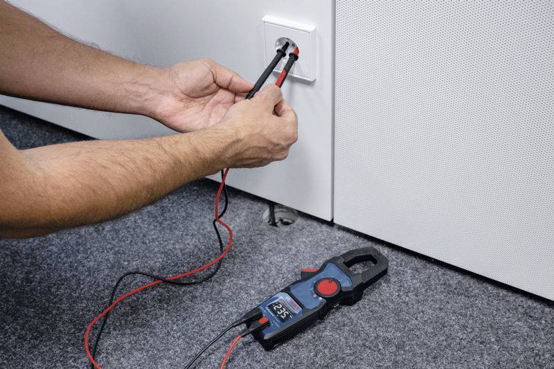 Eine Person misst die Spannung an einer Steckdose mit einem Multimeter. Das Display zeigt '225 V' an.