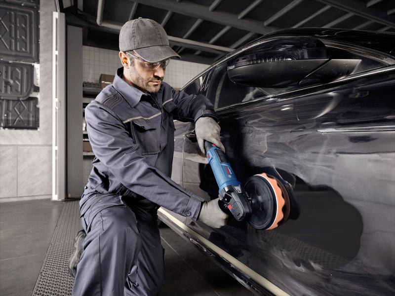 Ein Mechaniker poliert den Lack eines schwarzen Autos mit einer Poliermaschine in einer Werkstattumgebung.