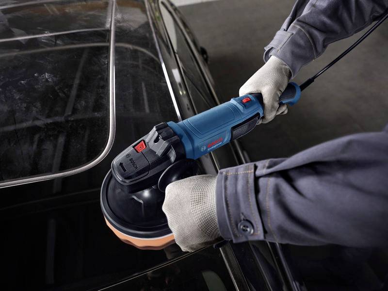 Eine Person in Arbeitskleidung poliert mit einer elektrischen Poliermaschine den Lack eines Autos.
