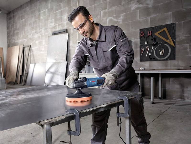 Ein Mann in Arbeitskleidung schleift mit einer elektrischen Poliermaschine eine Metallplatte in einer Werkstatt.