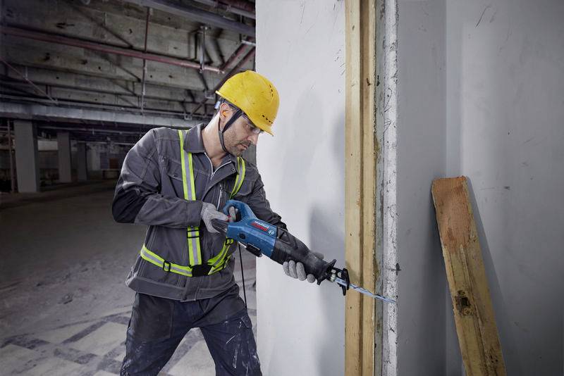 Ein Bauarbeiter mit gelbem Helm arbeitet mit einer Elektrosäge an einem Betonpfeiler in einer Baustelle. Sicherheitsweste sichtbar.