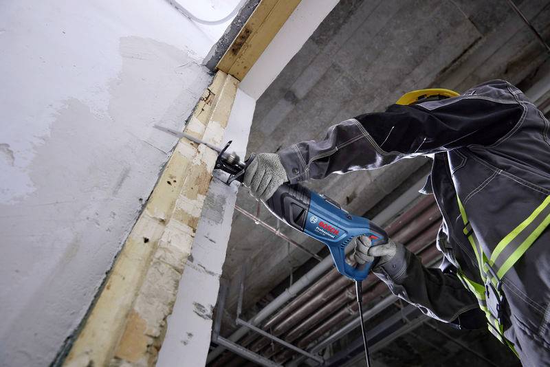 Eine Person trägt Schutzkleidung und verwendet eine elektrische Säge, um ein Stück Trockenbauwand unter einer Decke zu schneiden.