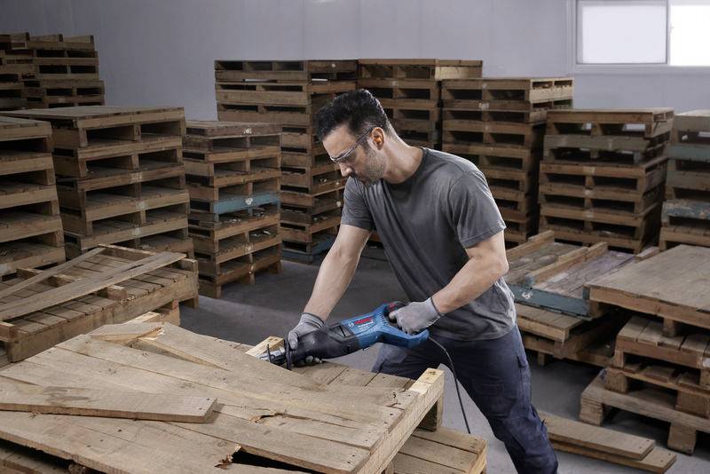 Ein Mann in Schutzbrille und Handschuhen zersägt eine Holzpalette mit einer Blaumann-Säge in einem Lagerraum voller gestapelter Paletten.
