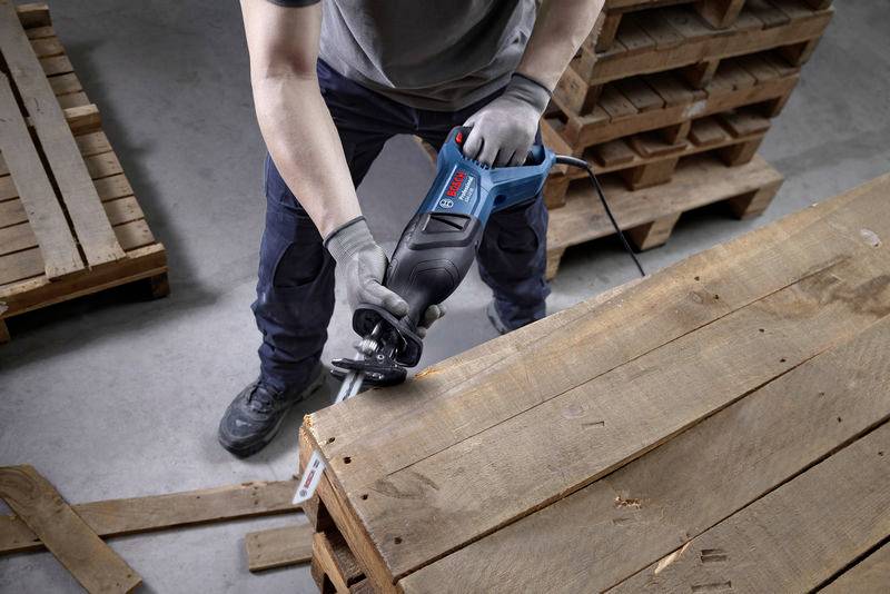 Eine Person verwendet eine elektrische Säge, um eine Holzpalette zu schneiden. Im Hintergrund befinden sich weitere Holzpaletten.