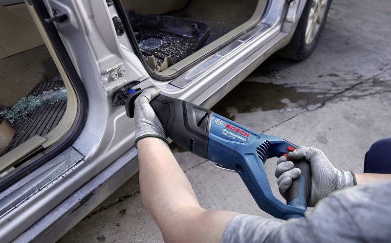 Person verwendet eine Elektrosäge, um eine Autotür zu entfernen. Sie trägt Sicherheitshandschuhe. Glasscherben liegen am Boden.