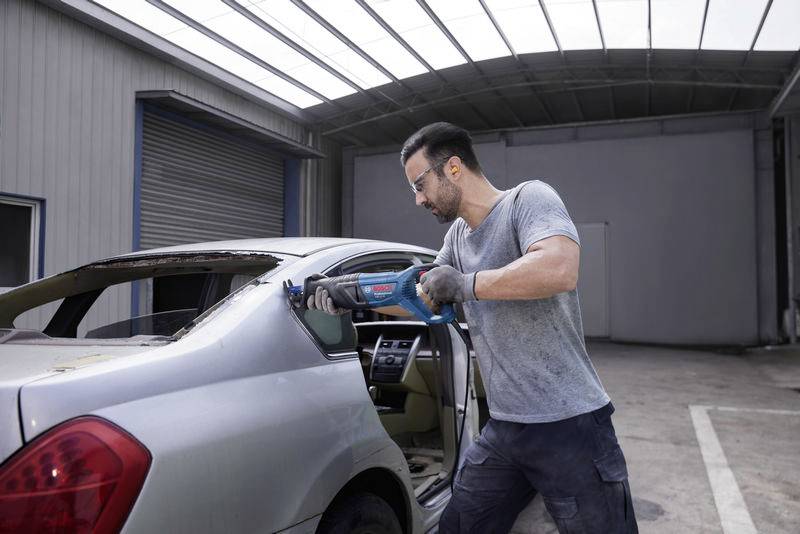 Ein Mann in Arbeitskleidung zersägt mit einer Elektrosäge das Heckfenster eines silbernen Autos in einer Werkstatt oder Garage.