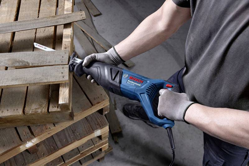 Eine Person sägt mit einer elektrischen Säge Holzpaletten in einer Werkstatt. Die Säge ist blau und hat ein Bosch-Logo.