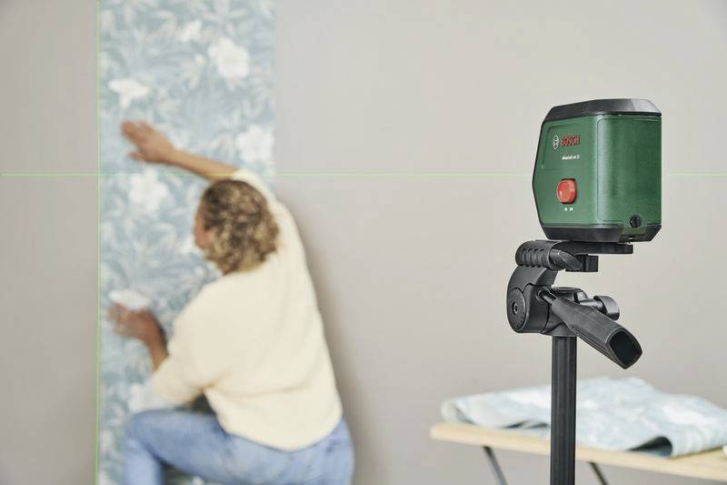 Eine Person tapeziert eine Wand entlang einer grünen Laserlinie, die von einem auf einem Stativ montierten Laserwerkzeug projiziert wird.