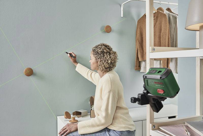 Eine Person benutzt ein Laserwerkzeug, um Linien an einer Wand zu markieren. Kleidung hängt im Schrank daneben.