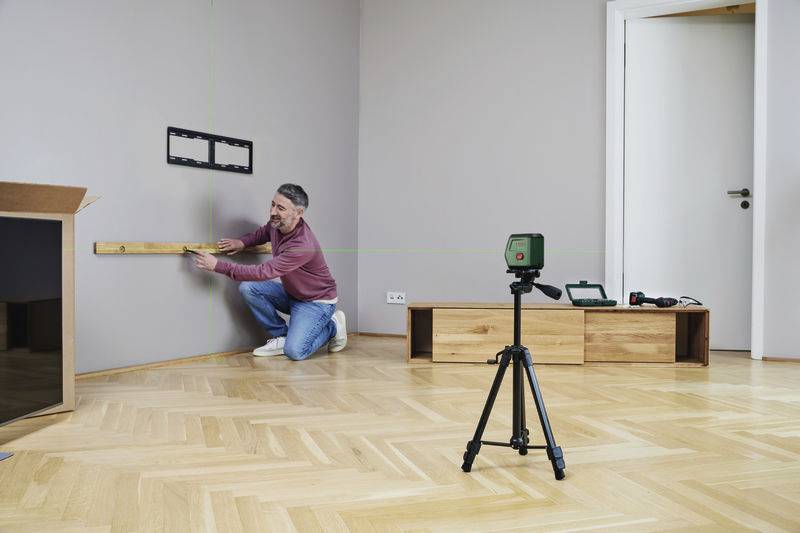 Eine Person montiert mit Wasserwaage und Laser ein Wandhalterung in einem Raum mit Holzboden, Kamera auf Stativ im Vordergrund.