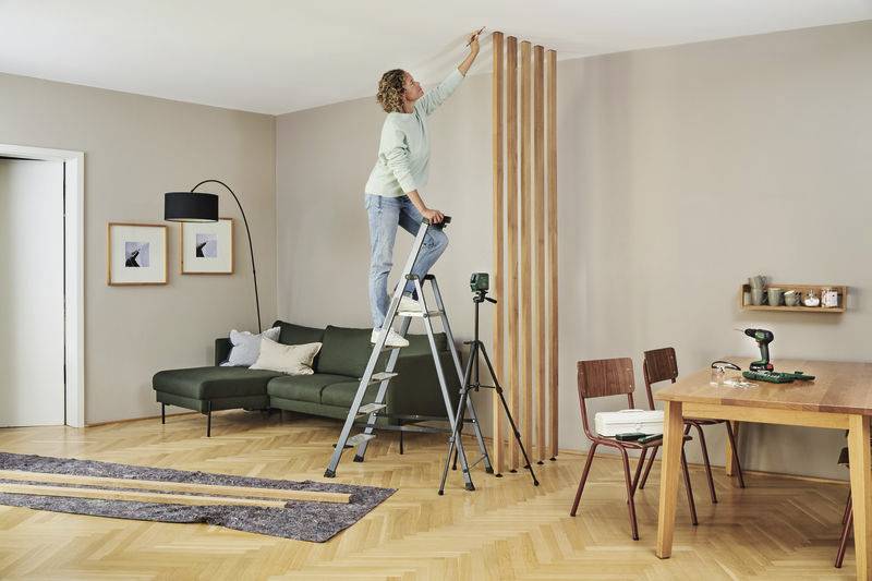 Eine Frau steht auf einer Leiter und streicht Holzlatten an einer Wand in einem Wohnzimmer. Neben ihr steht ein Stativ mit einem Lasergerät.